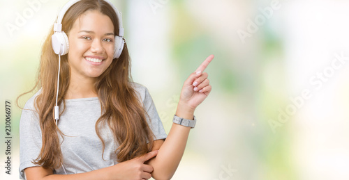 Young beautiful woman wearing headphones listening to music over isolated background with a big smile on face, pointing with hand and finger to the side looking at the camera.