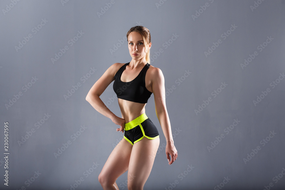 Obraz premium Lovely young woman bodybuilder posing in sports top and shorts on a gray background showing a beautiful sporty fit figure