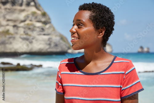 Horizontal shot of dark skinned teenager with curly short hair, piercing, smiles gently while looks aside with dreamy expression, wears casual t shirt poses over ocean view, blue sky wonderful scenery © Wayhome Studio