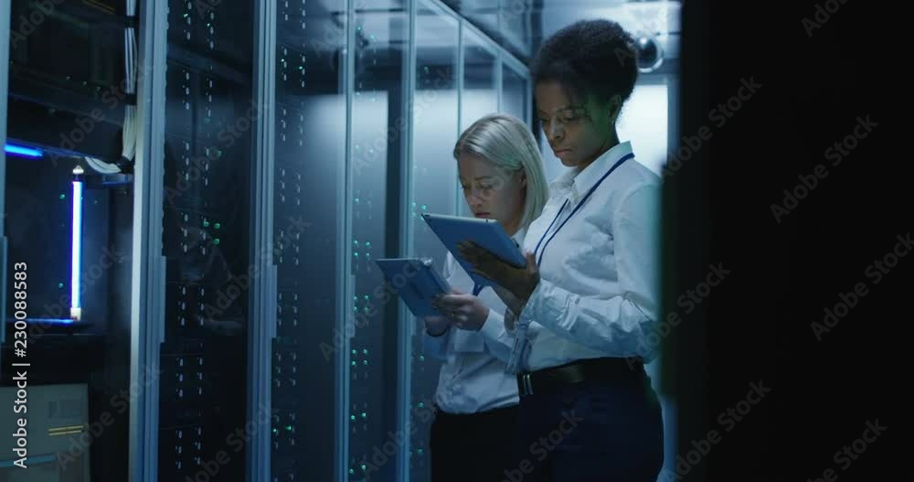 Medium shot of two women working in a data center with rows of server racks and checking the equipment and discussing their work