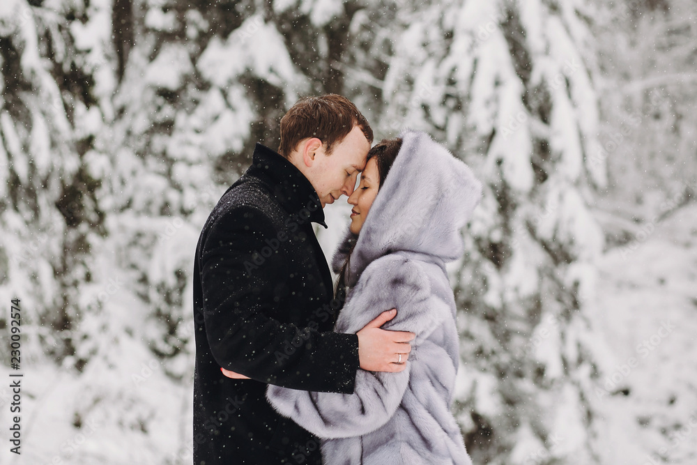 Stylish couple embracing in winter snowy mountains. Happy romantic man and woman in luxury clothes gently hugging at waterfall in snow. Holiday getaway together Stylish couple embracing in winter snowy mountains. Happy romantic man and woman in luxury clothes gently hugging at waterfall in snow. Holiday getaway together