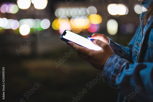 Woman pointing finger on blank screen smartphone on background bokeh light in night atmospheric city. hipster using in hands clean gadget mobile phone closeup, mockup street, online wifi internet