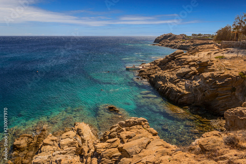 Summertime: Mykonos island, Greece. Beautiful scenery of rocky coast: azure waters of the Aegean Sea, cliff, rocks and stones. Sunny with blue sky and crystal clear water. 