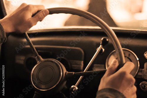driving a vintage car; only the driver’s hand on the steering wheel are visible, the dashboard is blurred; stylized as an old sepia photo with dust and noise..