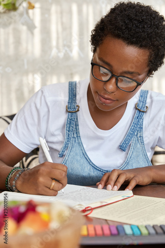 Dark skinned hipster in eyewear, writes in notebook, does homework , sits at desk top, works on diploma paper. Female freelance with Afro haircut rewrites indormation in diary, holds pen, sits at cafe photo