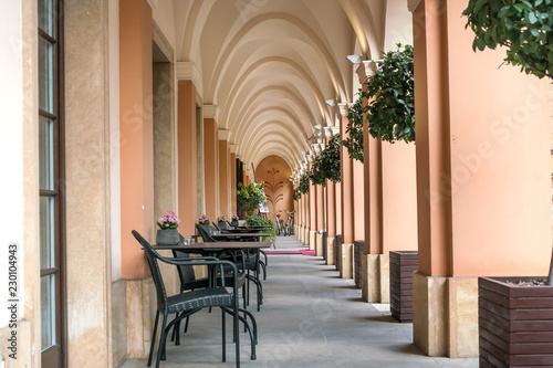 part of the street restaurant cafe under the arch with columns, flowers and greenery surroundings, vintage, empty tables and chairs along the long wall