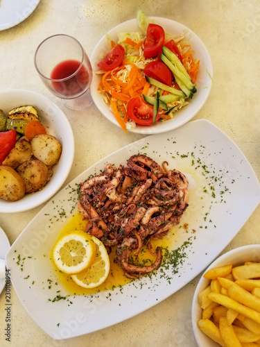 Fried octopus with lemon and spices, vegetables and french fries on white plate with glass of red wine in summer street cafe in Marsaxlokk, Malta