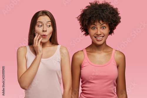 Lovely woman looks surprisingly at dark skinned cheerful friend, feels envy about something, dressed in casual clothes, poses against pink background. Multiethnic colleagues express different emotions