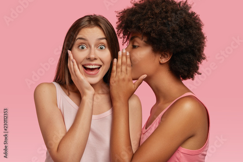 Young black woman whispers secret to her Caucasian friend, gossip together and spread rumours. Emotional European girl feels amazed to hear confidential information from companion. Secrecy, gossiping photo