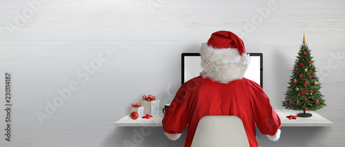 Santa Claus working on a computer in his office during Christmas holidays. Empty space on wall for text.