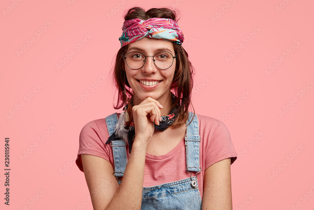 Studio shot of attractive hippie female holds chin, has broad smile, wears round spectacles, fashionable clothes, has pleasant talk with hipster guy, express positiveness, isolated on pink wall Studio shot of attractive hippie female holds chin, has broad smile, wears round spectacles, fashionable clothes, has pleasant talk with hipster guy, express positiveness, isolated on pink wall