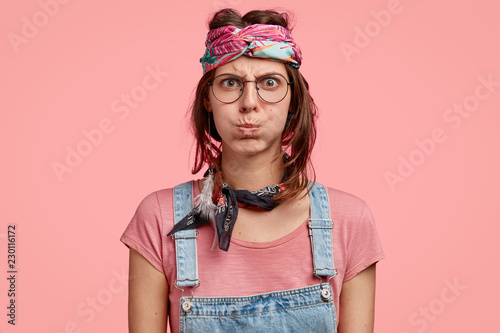 Grumpy sullen hippie woman blows cheeks with dispasure, has angry facial expression, wears headband and denim overalls, has quarrel with compnion, considers herself to be right, isolated on pink photo