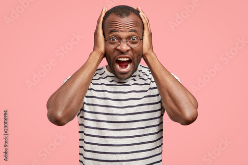 Photo of depressed angry dark skinned man being under pressure, keeeps hands on head, exclaims angrily, dressed in striped t shirt, isolated over pink background. People and negative emotions