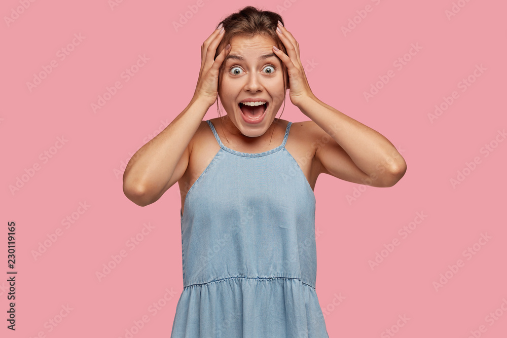 Indoor shot of amazed young European woman exclaims very loudly, keeps hands on head, stares with bugged green eyes, models against pink studio wall. People, excitement and happiness concept. Indoor shot of amazed young European woman exclaims very loudly, keeps hands on head, stares with bugged green eyes, models against pink studio wall. People, excitement and happiness concept.