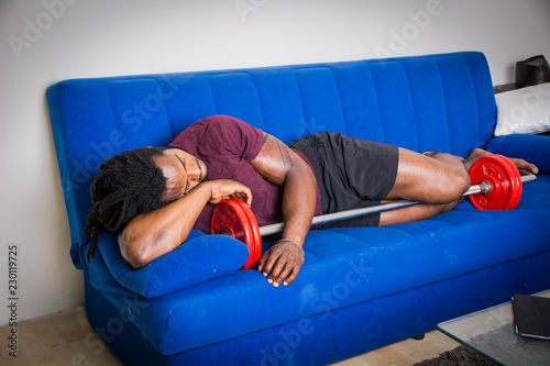 Young strong black male sleeping on couch embracing barbell. photo