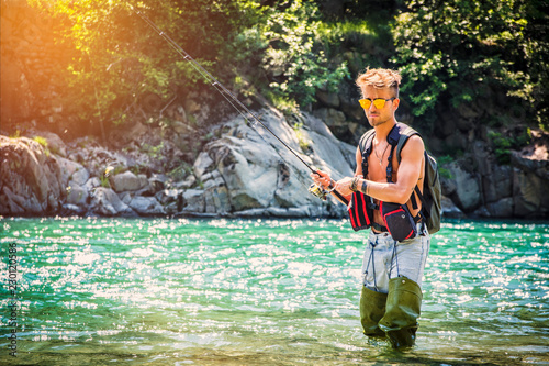 Young handsome male tourist standing and fishing in river in sunny day with rod