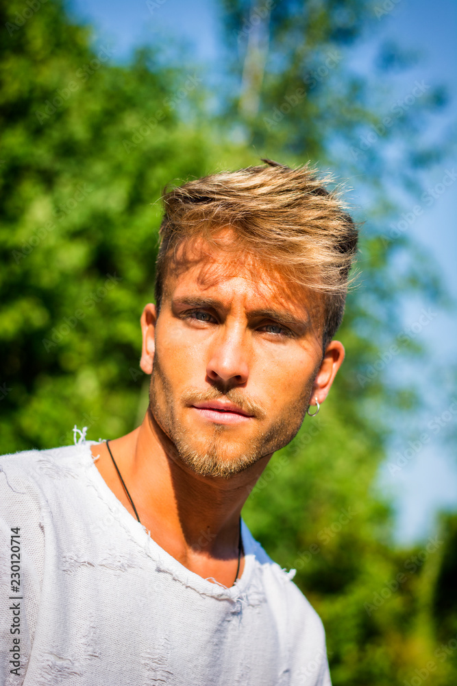 Blond, blue eyed young man outdoor in the sun, thinking, looking at camera, with green trees behind him Blond, blue eyed young man outdoor in the sun, thinking, looking at camera, with green trees behind him