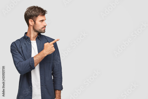 Stylish unshaven young man with dark bristle, attracts your attention to something, points with index finger at blank space, shows free space for your advertising content, stands sideways indoor photo