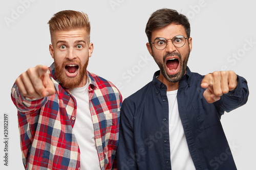 Peevish angry men with thick stubbles indicate at camera with index fingers, makes you guilty, shout angrily at someone, have annoyed expressions, model against white background. Just look forward
