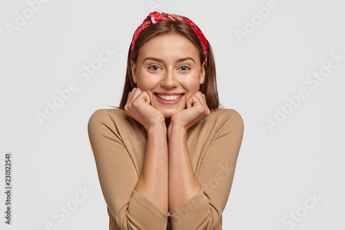 Horizontal shot of pretty Caucasian woman holds chin with both hands, looks happily at camera, wears casual beige swater and headband, chuckls from happiness, models against white background photo