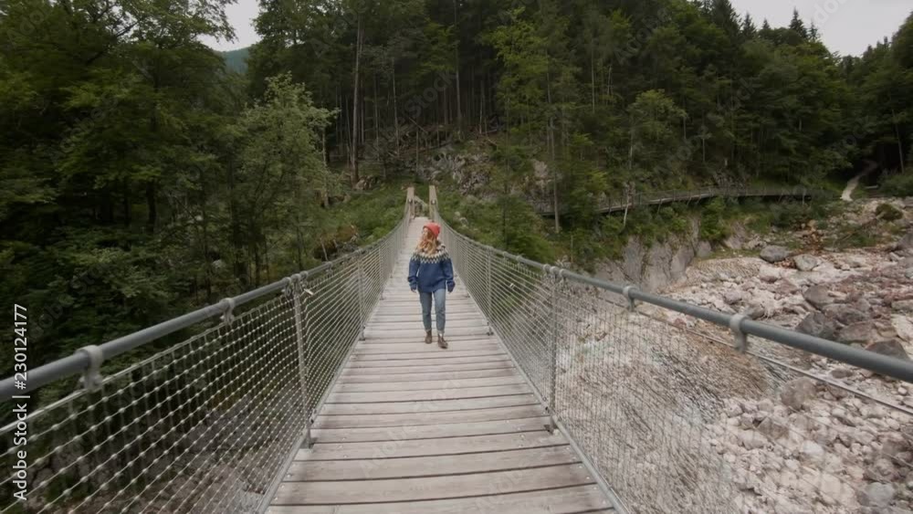 Young female traveler or explorer walks confidently on small wooden bridge hanging over mountain river, wears authentic wool sweater, lives nomad travel blogger lifestyle Young female traveler or explorer walks confidently on small wooden bridge hanging over mountain river, wears authentic wool sweater, lives nomad travel blogger lifestyle