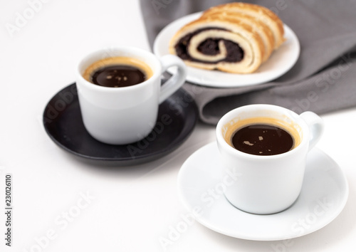 White cups of coffee on black saucer pad with poppy seed cake in behind.