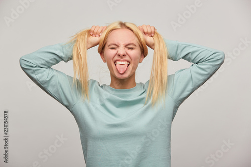 Cool young blonde girl with long hair grimacing on camera, showing tongue, eyes closed, has fun, makes two tails on her head, wearing blue casual sweatshirt,i solated over grey background photo