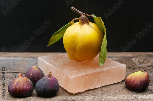 A single yellow quince with leaves and stems on a Himalayan salt block with figs on a rustic table top