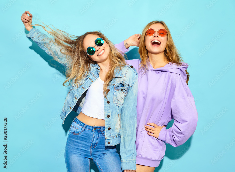 Two young beautiful blond smiling hipster girls in trendy summer clothes. Sexy carefree women posing near blue wall in sunglasses. Positive models going crazy Two young beautiful blond smiling hipster girls in trendy summer clothes. Sexy carefree women posing near blue wall in sunglasses. Positive models going crazy
