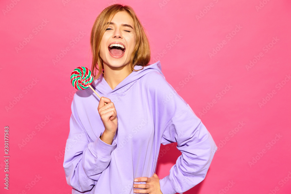 Portrait of young beautiful smiling hipster girl in trendy summer hoodie. Sexy carefree woman posing near pink wall. Positive model with lollipop Portrait of young beautiful smiling hipster girl in trendy summer hoodie. Sexy carefree woman posing near pink wall. Positive model with lollipop