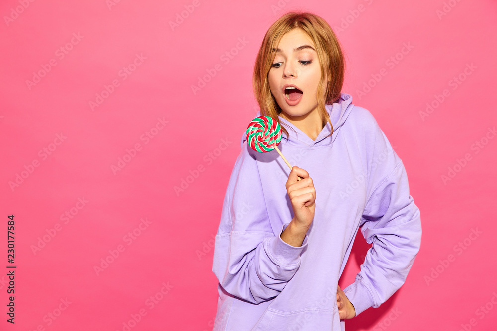 Portrait of young beautiful smiling hipster girl in trendy summer hoodie. Sexy carefree woman posing near pink wall. Positive model with lollipop. Portrait of young beautiful smiling hipster girl in trendy summer hoodie. Sexy carefree woman posing near pink wall. Positive model with lollipop.