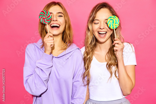 Two young beautiful smiling blond hipster girls in trendy summer clothes. Carefree hot women posing near pink wall. Positive models cover eyes by lollipop