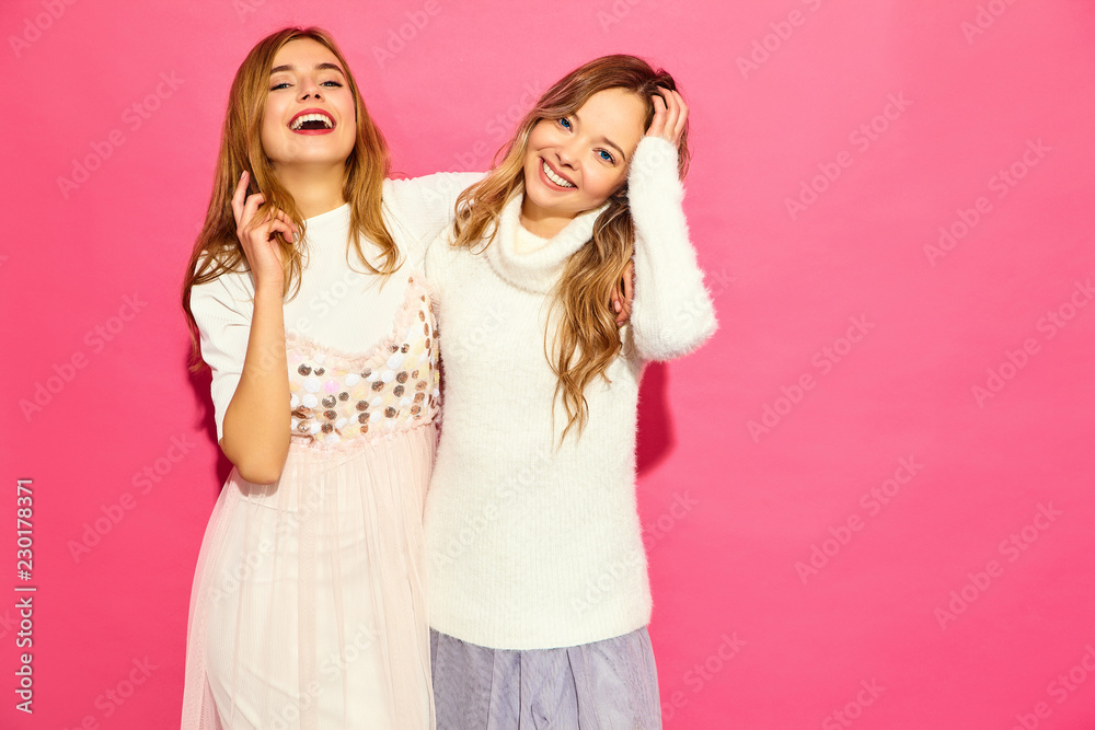 Two young beautiful smiling hipster girls in trendy summer white clothes. Sexy carefree women posing near pink wall. Positive models