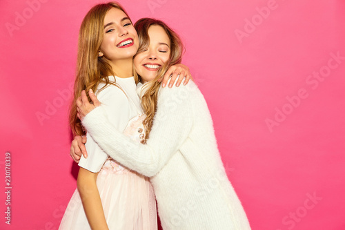 Two young beautiful smiling hipster girls in trendy summer white clothes. Sexy carefree women posing near pink wall. Positive models hugging