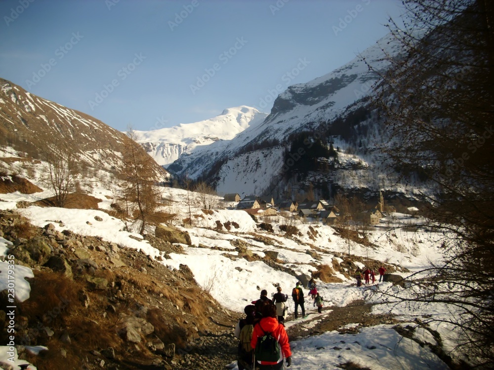 Groupe de randonneurs en raquettes dans une vallée alpine marchant vers un village de montagne. Sommets enneigés et ciel bleu pur en arrière plan. 