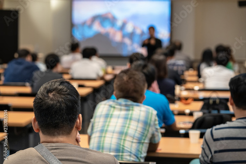 Speaker on the stage in front of the room with Rear view of Audience in put hand up acton for answer the question in the meeting or seminar meeting, business and education concept