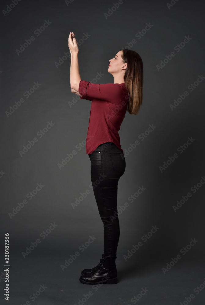 full length portrait of brunette girl wearing  red shirt and leather pants. standing pose , on grey studio background.