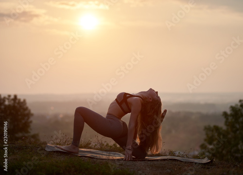 Silhouette of attractive slender barefooted girl with beautiful long golden hair in summer training outfit in complicated gymnastic yoga posture on grassy hill under bright pink misty sky at sunset.