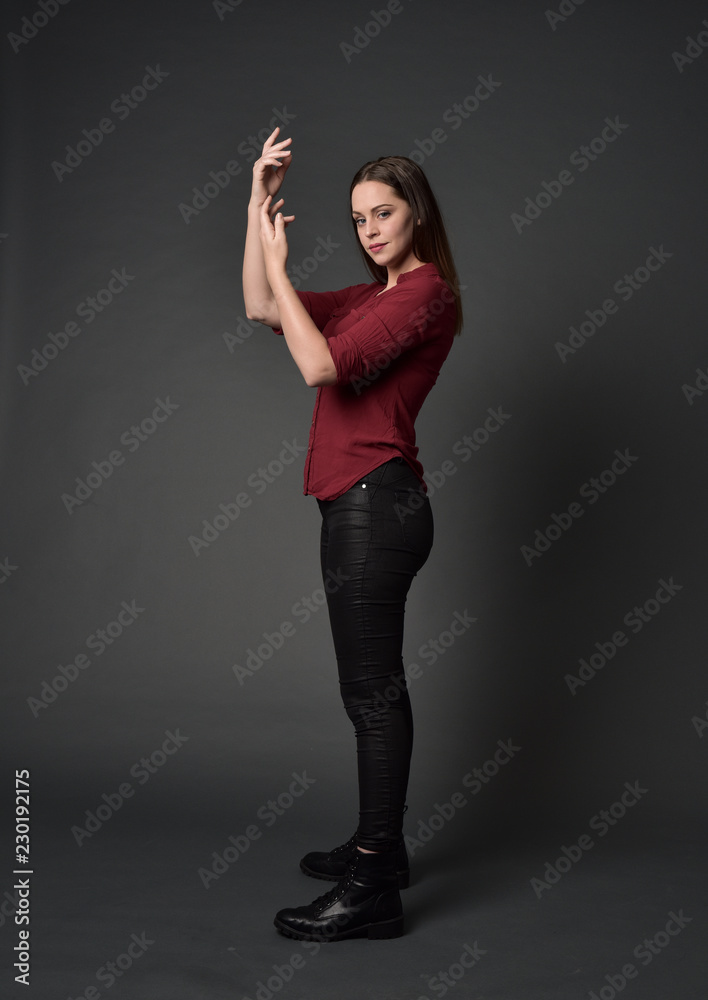 full length portrait of brunette girl wearing  red shirt and leather pants. standing pose , on grey studio background.