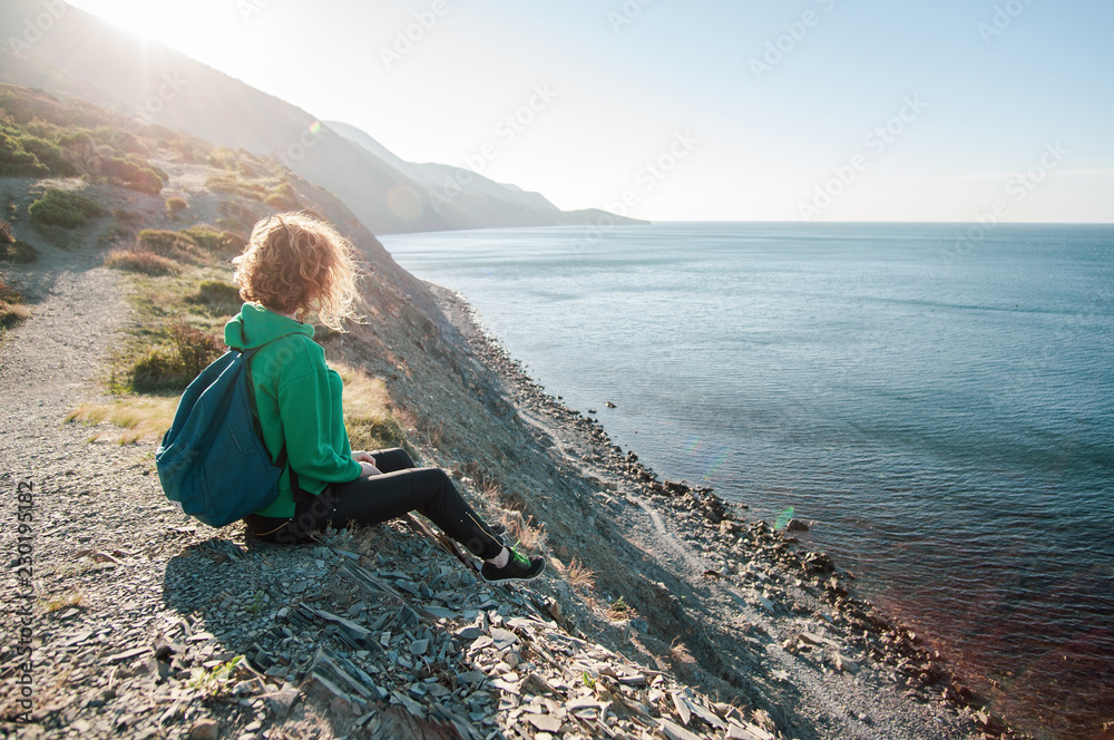 The girl-traveler enjoys the sea view sitting on the edge of the cliff, side view