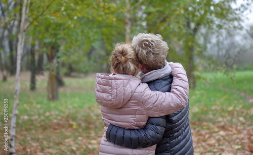 mother and daughter in park, give hug each other, from their back, both blond hair, autumn, background in soft focus.