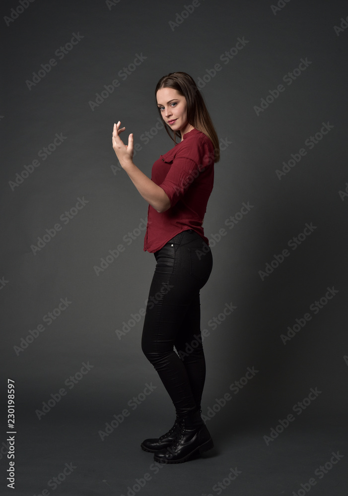 full length portrait of brunette girl wearing  red shirt and leather pants. standing pose , on grey studio background.