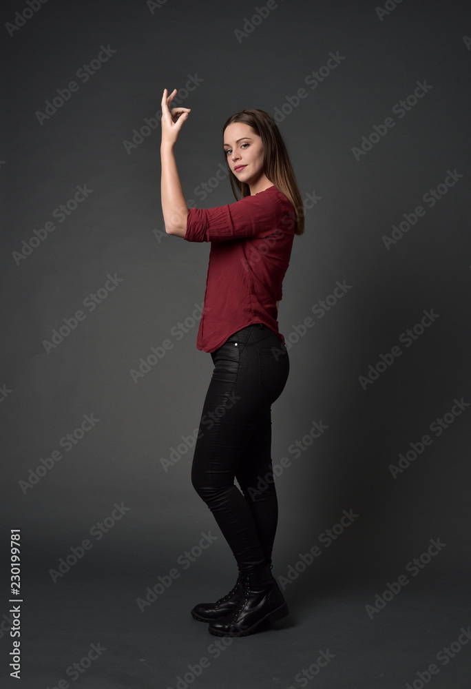 full length portrait of brunette girl wearing  red shirt and leather pants. standing pose , on grey studio background.