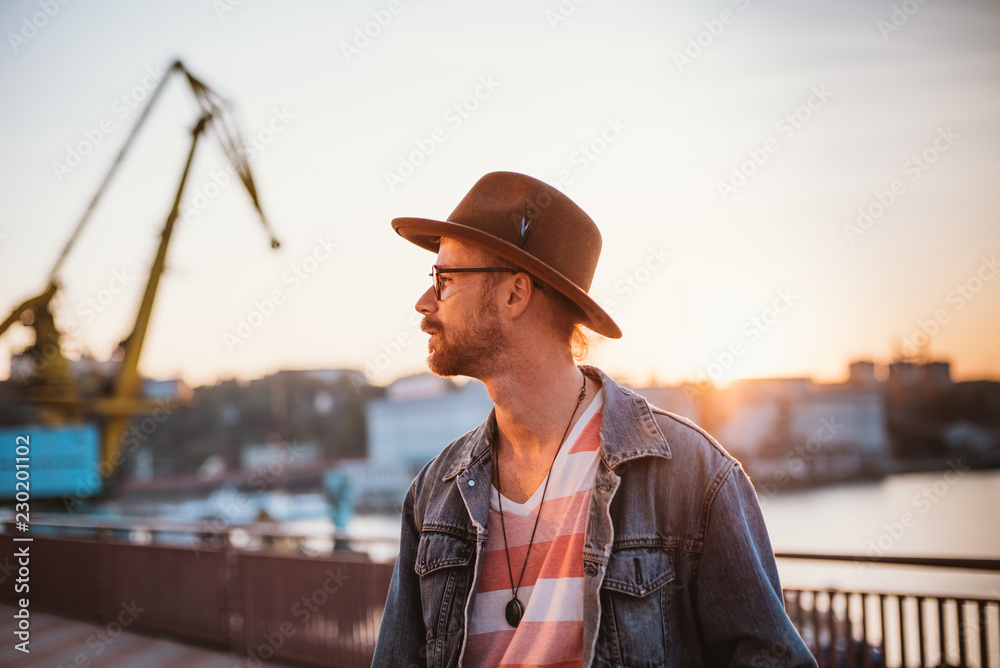Young stylish hipster in round glasses and hat on sea port background during sunset Young stylish hipster in round glasses and hat on sea port background during sunset
