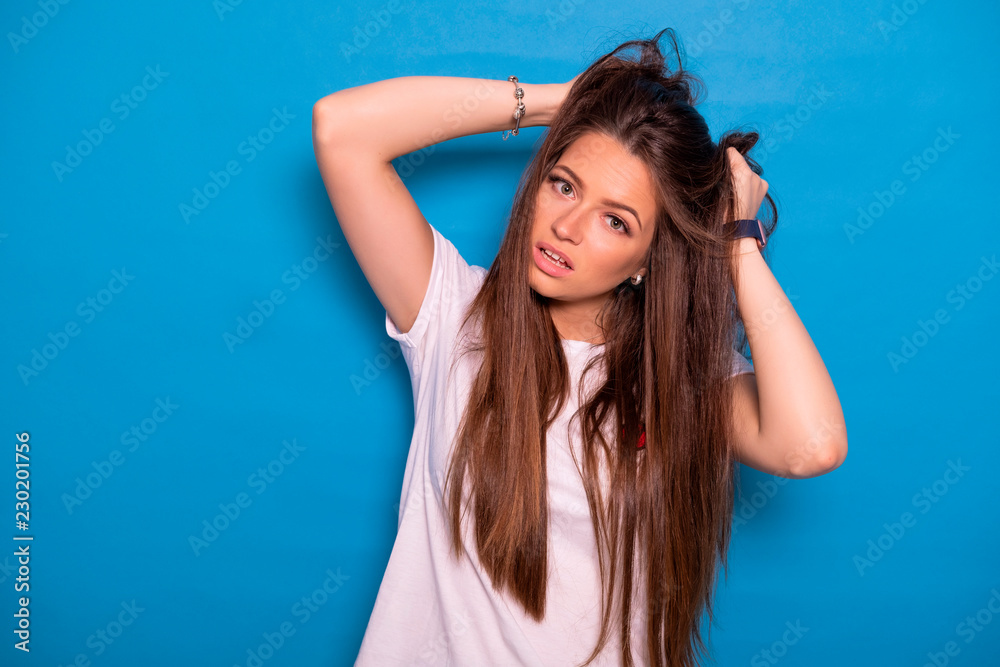 Cute brunette woman with long hair posing in white t-shirt on a blue background. Emotional portrait. She tries to tear her hair