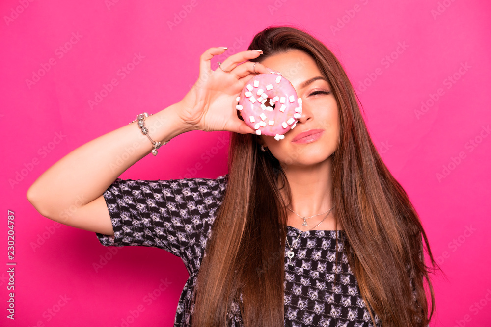 Sexy model with long brunette hair on a pink background. Emotional portrait. She eating donut
