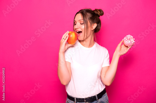 Sexy model with stylish hairstyle in white t-shirt on a pink background. Emotional portrait. She shocked, amazed, tying to choose between good and bad food: apple or donut?