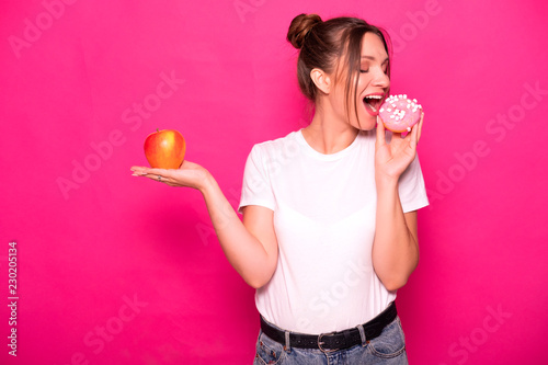 Sexy model with stylish hairstyle in white t-shirt on a pink background. Emotional portrait. She shocked  amazed  tying to choose between good and bad food  apple or donut 