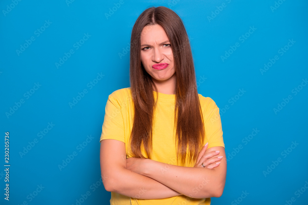 Cute brunette woman with long hair posing in yellow t-shirt on a blue background. Emotional portrait. She shows her disgust