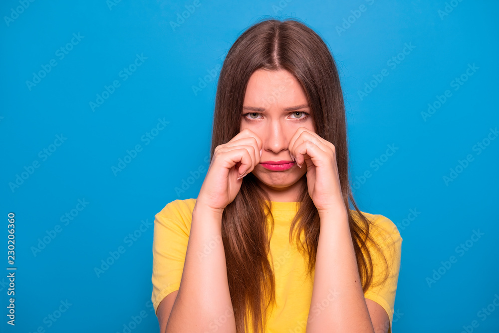 Cute brunette woman with long hair posing in yellow t-shirt on a blue background. Emotional portrait. She sad, cry Cute brunette woman with long hair posing in yellow t-shirt on a blue background. Emotional portrait. She sad, cry